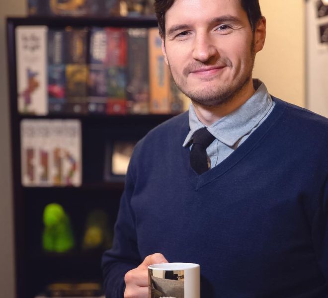 male therapist holding a coffee mug, wearing a blue sweater poses smiling in his office doorway with a bookcase in the background