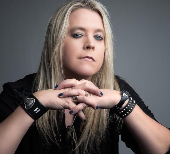 blonde woman dressed in black, poses with her elbows on the table in front of her, and a grey background