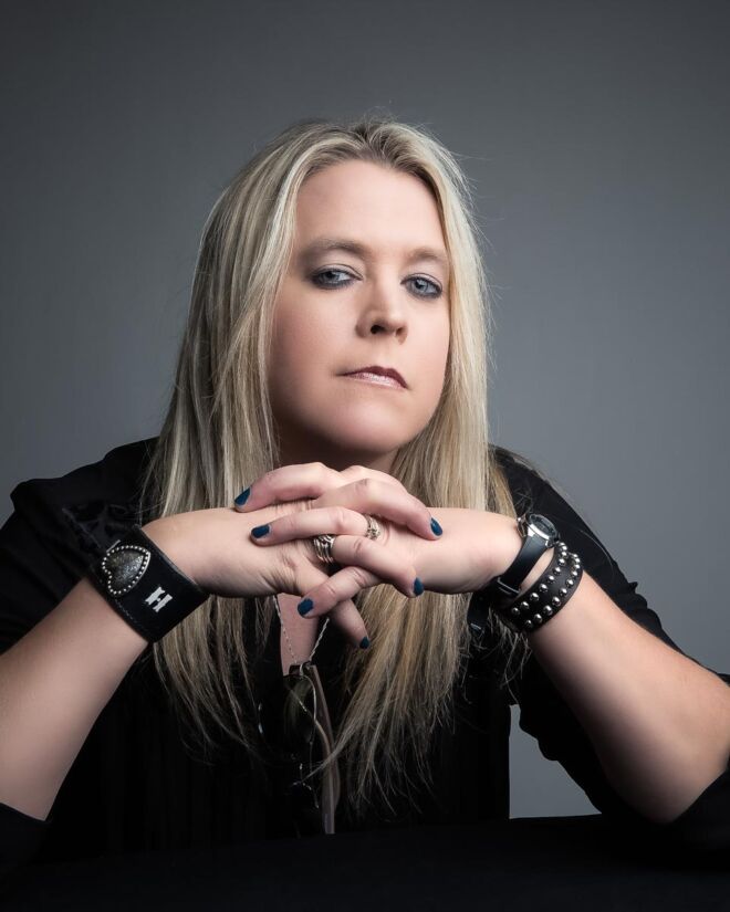 blonde woman dressed in black, poses with her elbows on the table in front of her, and a grey background