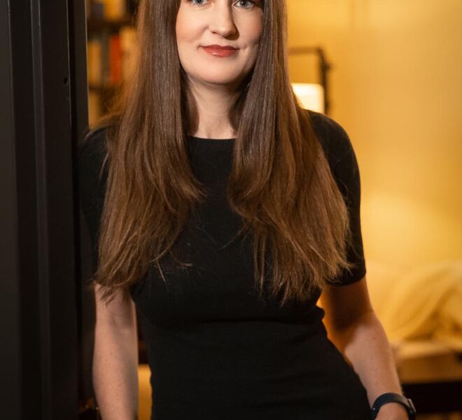 Attractive young female therapist in a black top and black jeans poses smiling in her office doorway with her hands in her pockets.