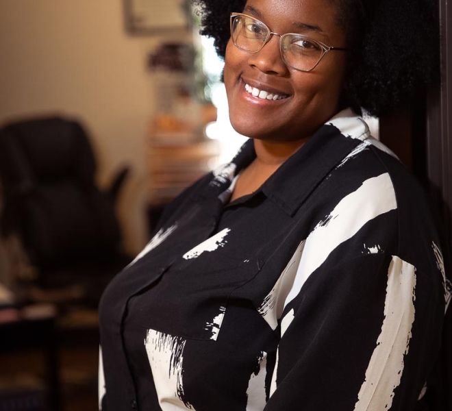 Young african american female in glasses and a black and white top poses smiling in her office doorway