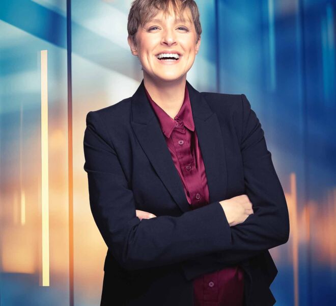 blonde short haired business woman laughs in front of a blue and orange urban window background, wearing a burgundy top and a blue blazer