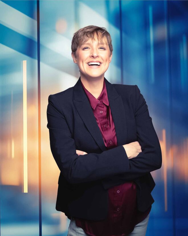 blonde short haired business woman laughs in front of a blue and orange urban window background, wearing a burgundy top and a blue blazer