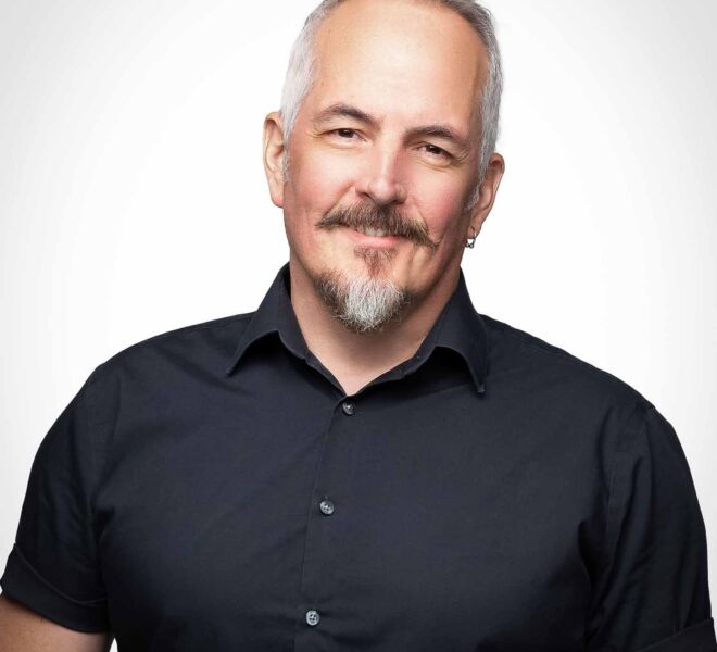handsome middle aged man with salt and pepper hair, mustache and beard poses smiling in a black button up short sleeve shirt for headshots in Tulsa
