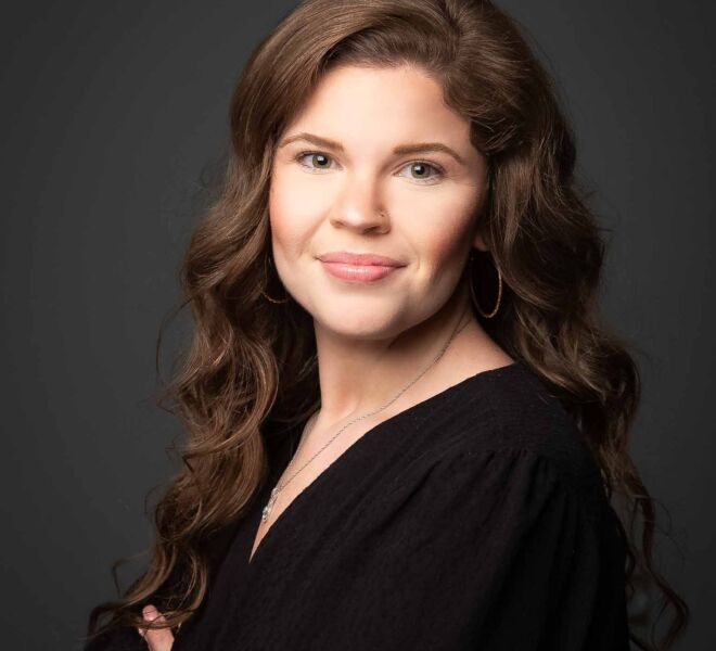 attractive professional woman with wavy brown hair and hazel eyes crosses her arms and smiles on a dark grey background for her headshot at tulsa hard rock casino
