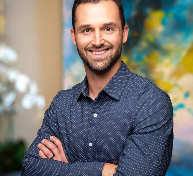 Young businessman with a beard smiles for headshot. Wearing a blue button down shirt with his arms crossed in an office setting