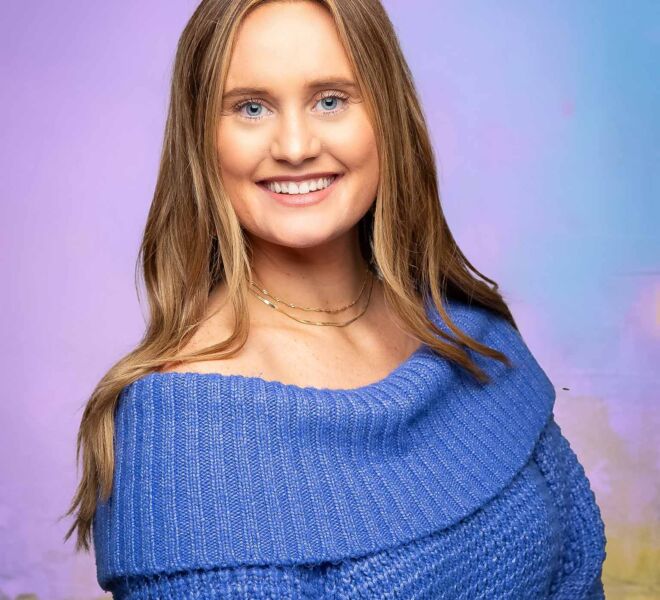 Pretty blue eyed, blonde woman in a blue sweater poses smiling for a headshot in front of a pink gold blue and purple background