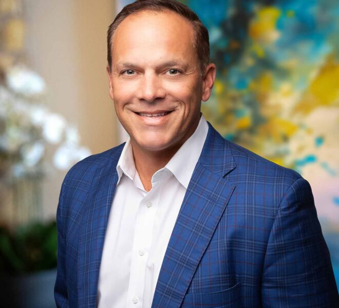 businessman poses smiling in an office setting, wearing a blue tartan patterned jacket and white shirt