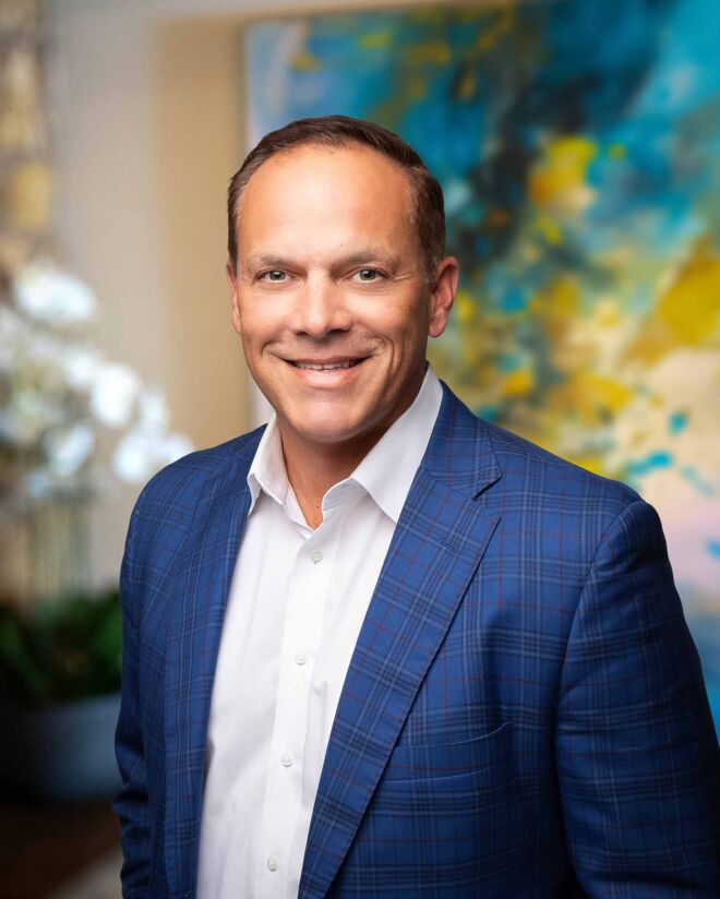 businessman poses smiling in an office setting, wearing a blue tartan patterned jacket and white shirt