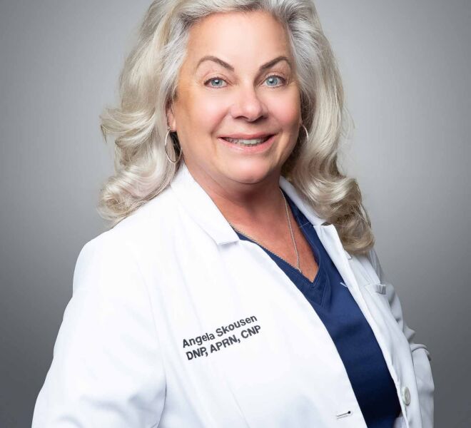 white haired female doctor wearing a white lab coat and a blue top poses smiling in front of a grey background