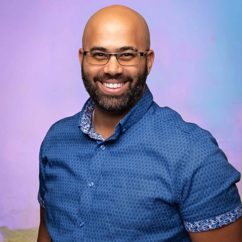 tulsa headshots_519 man in blue short sleeve button up shirt with glasses, smiling in front of a purple background
