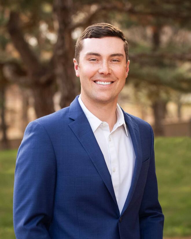 young businessman poses outdoors in a blue jacket and white shirt, smiling for a Tulsa headshot