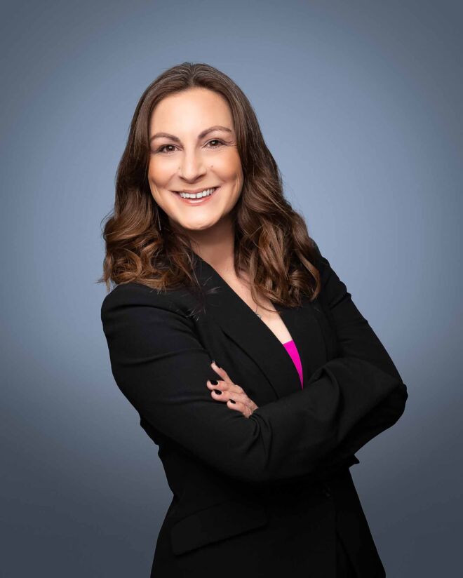 Female tulsa realtor crosses her arms and smiles in a black suit with a pink top on a grey background