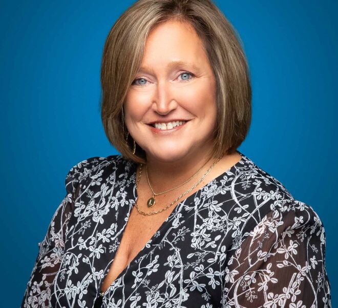 Woman with blue eyes and short hair wearing a black and white dress smiles for a headshot portrait while posing on a blue background
