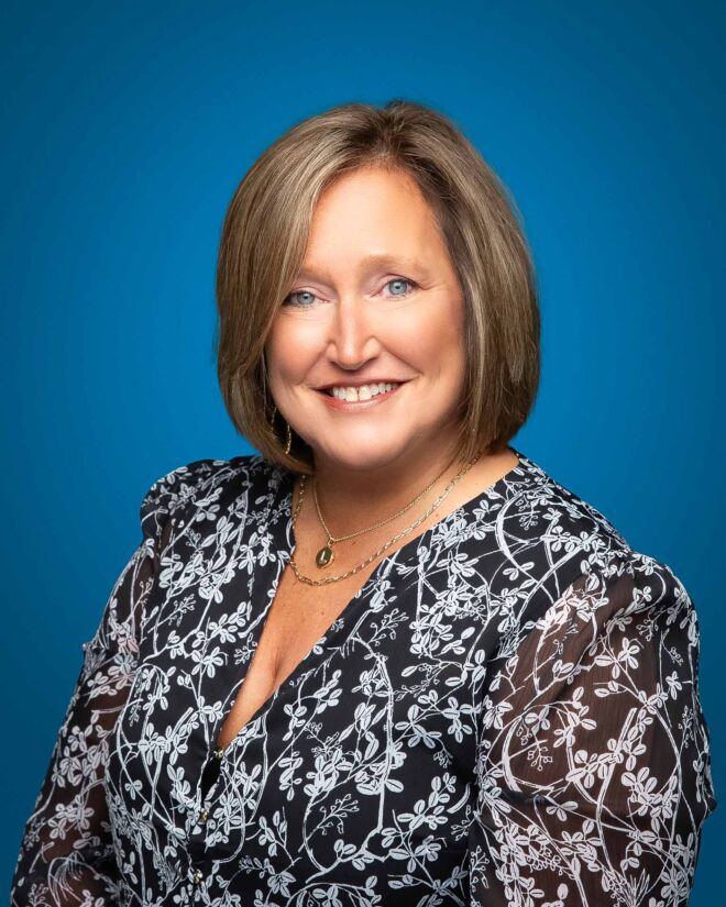 Woman with blue eyes and short hair wearing a black and white dress smiles for a headshot portrait while posing on a blue background