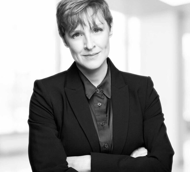 Black and white headshot photo of a businesswoman, Slightly smiling, arms crossed, short hair in a brightly lit office setting