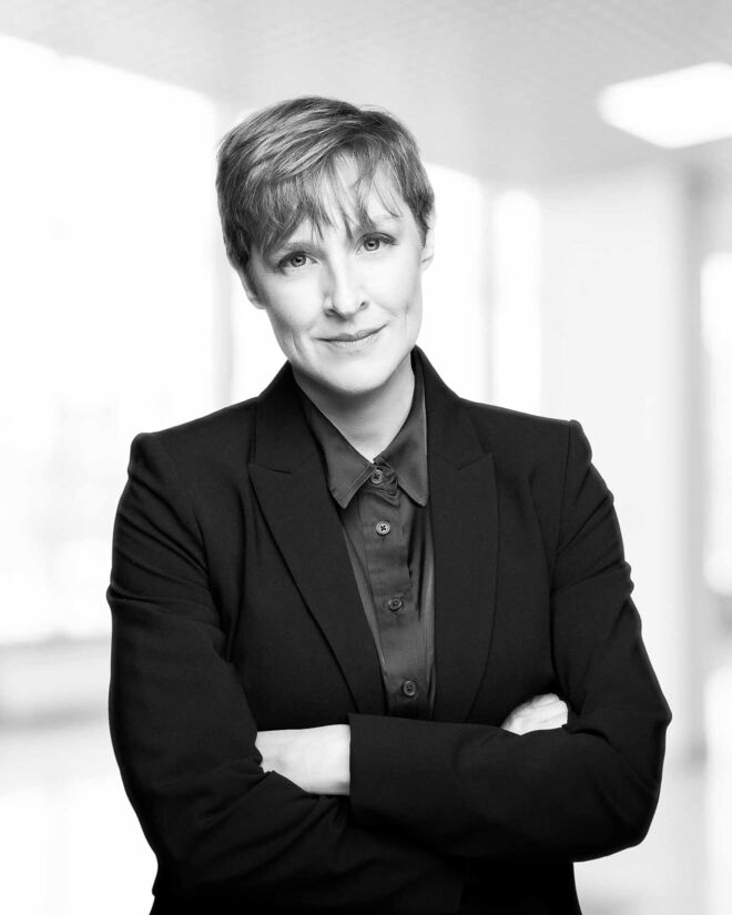 Black and white headshot photo of a businesswoman, Slightly smiling, arms crossed, short hair in a brightly lit office setting