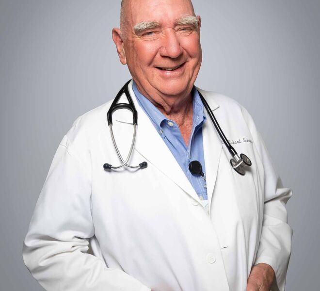 Older male doctor with bushy eyebrows laughs and smiles, wearing a white lab coat and blue shirt in front of a grey background