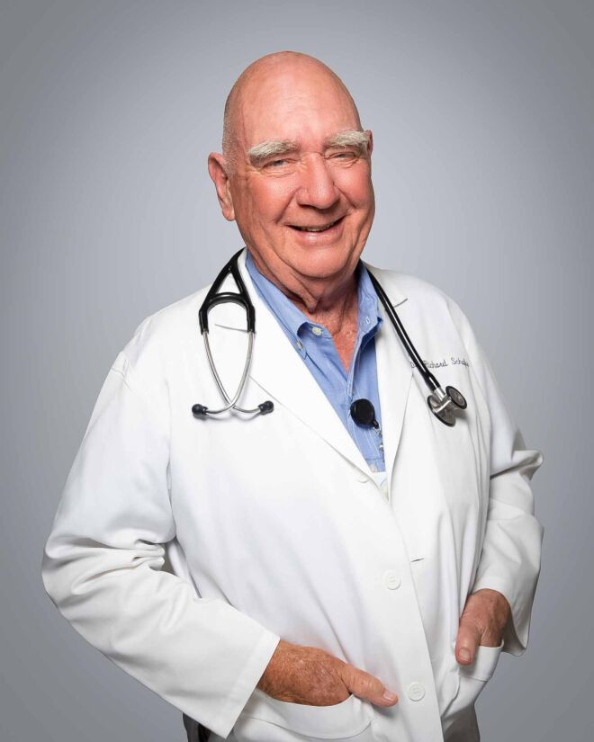 Older male doctor with bushy eyebrows laughs and smiles, wearing a white lab coat and blue shirt in front of a grey background