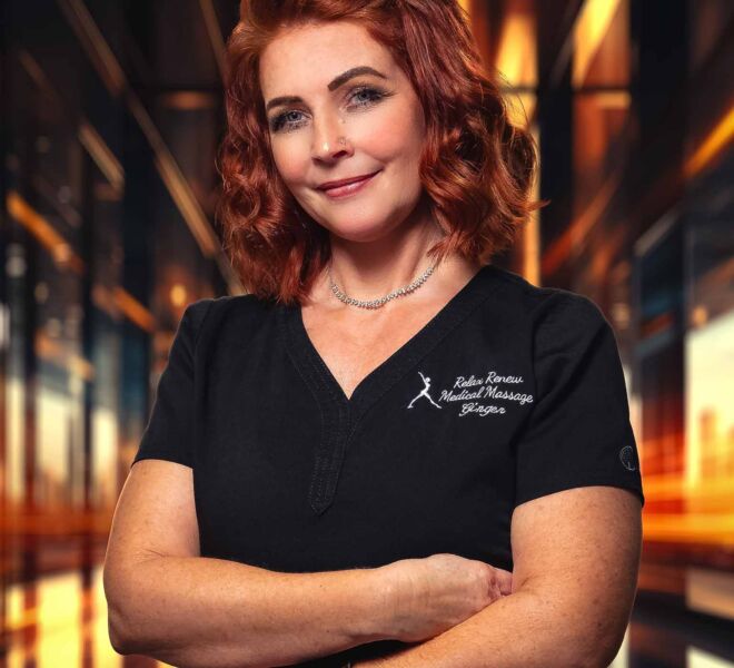red haired medical massage therapist in black scrubs stands smiling with arms crossed in an orange and black backlist office setting