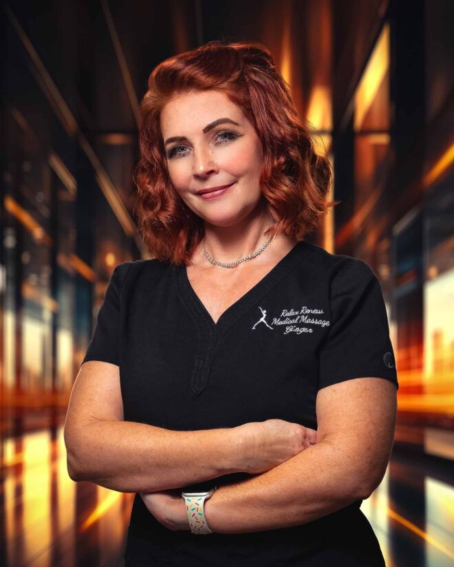 red haired medical massage therapist in black scrubs stands smiling with arms crossed in an orange and black backlist office setting