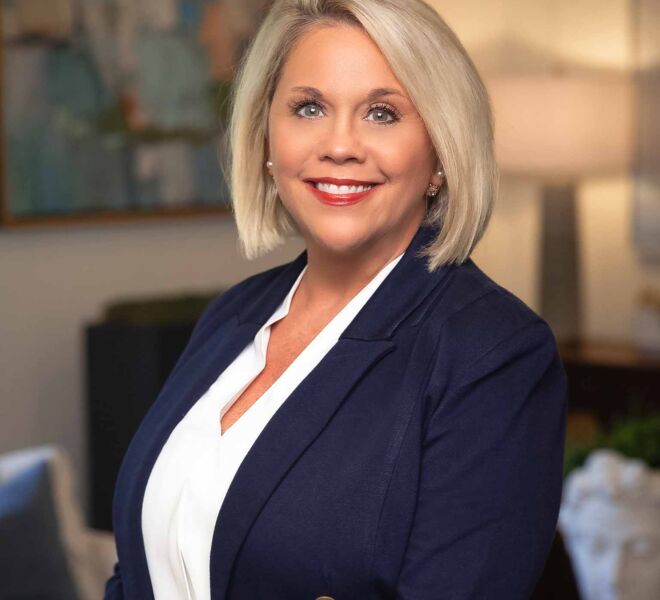 Blonde businesswoman smiles in an office setting, wearing a blue jacket and white top.
