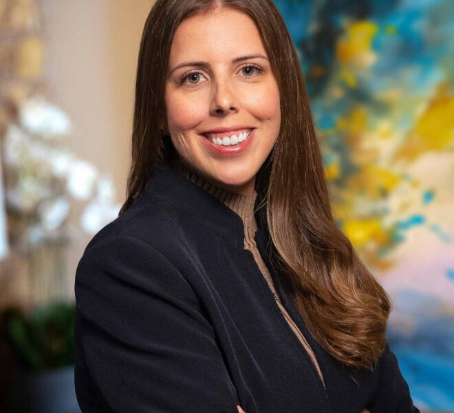 smiling young woman in a blue jacket and brown sweater poses with arms crossed in front of a blue office background.
