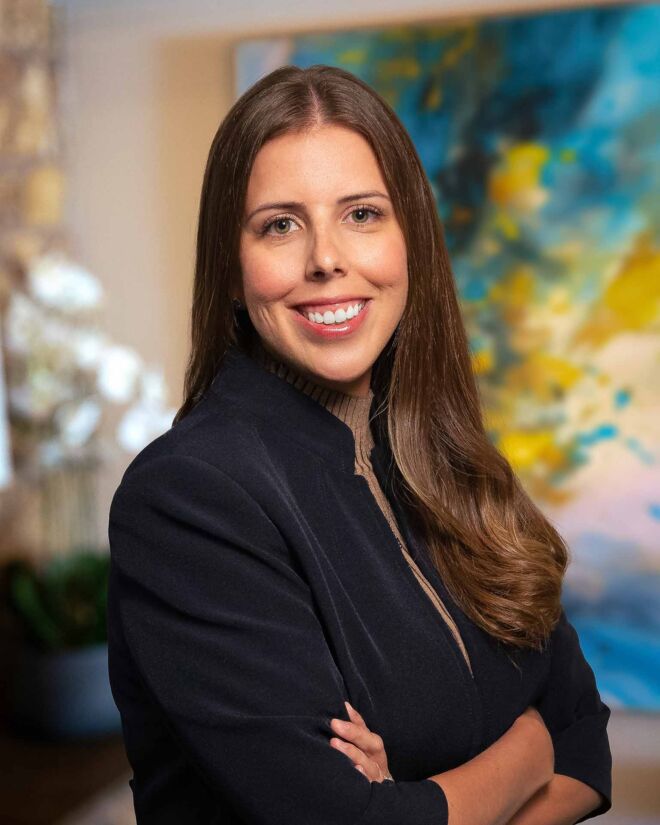 smiling young woman in a blue jacket and brown sweater poses with arms crossed in front of a blue office background.