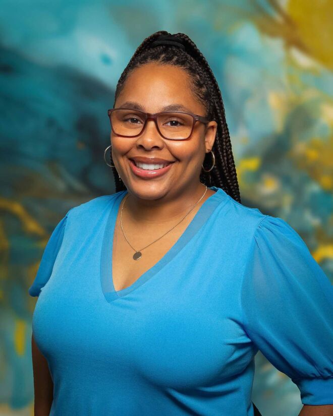 African American businesswoman in glasses and a blue top, smiles and poses for headshots in front of a blue and gold background