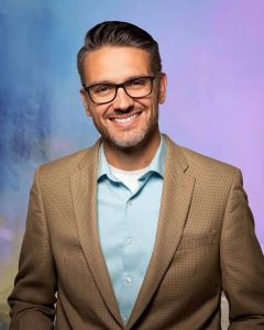 Man in glasses, brown sports coat and blue shirt, smiling in front of a purple and blue studio background.