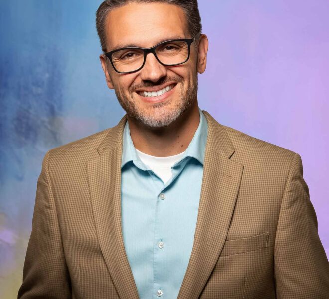 Man in glasses, brown sports coat and blue shirt, smiling in front of a purple and blue studio background.