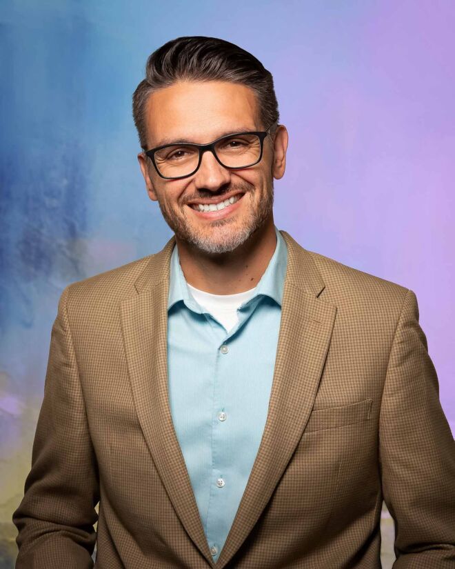 Man in glasses, brown sports coat and blue shirt, smiling in front of a purple and blue studio background.