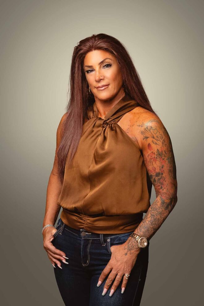 Attractive female tulsa realtor posing on a grey background wearing blue jeans and a copper colored top with her thumbs in her pockets