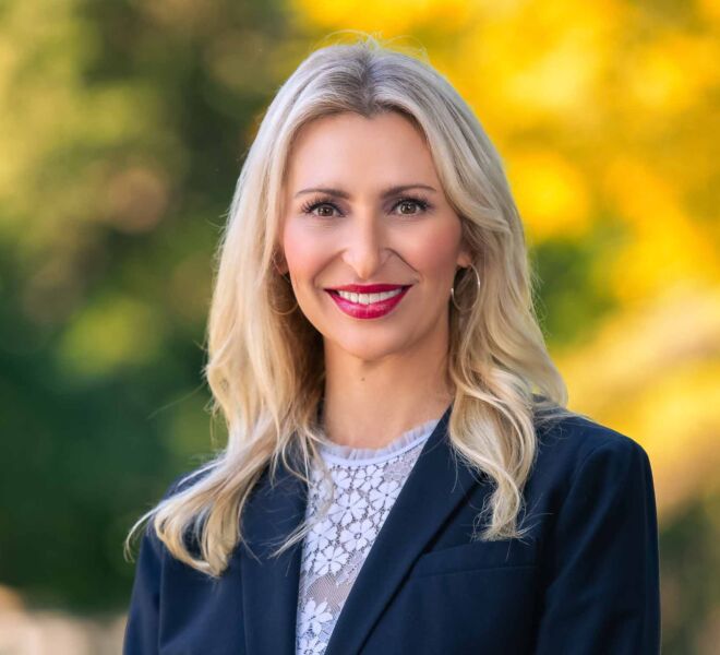 Attractive blonde businesswoman in a navy blue blazer and white top, smiling in a shallow depth of field headshot outdoors