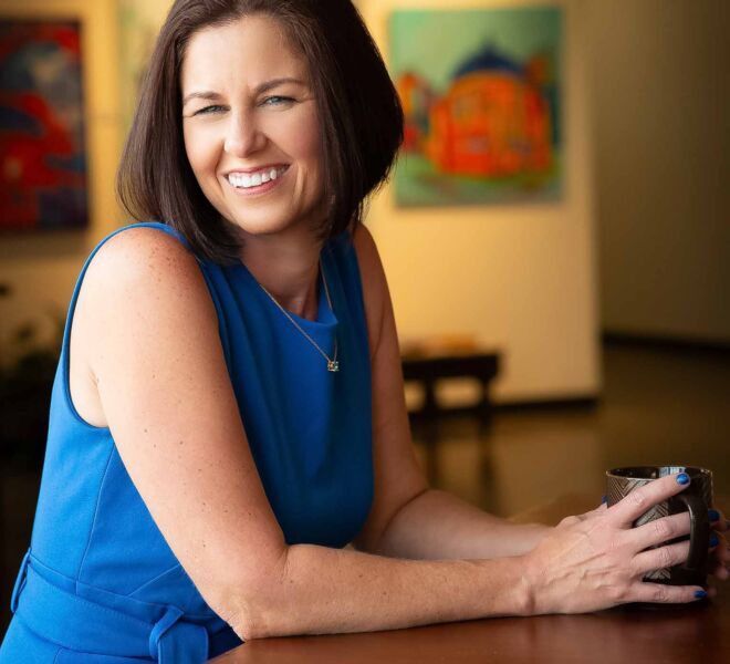 Happy woman in a blue top, having coffee at a table with paintings on the wall behind her