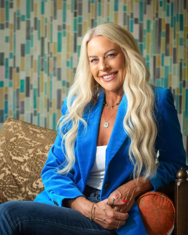 Beautiful blonde woman smiling on a couch, wearing a blue blazer in front of a colorful background.