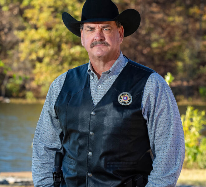 Wagner County Sheriff in cowboy hat, black leather vest and blue shirt posing in front of a lake for headshots