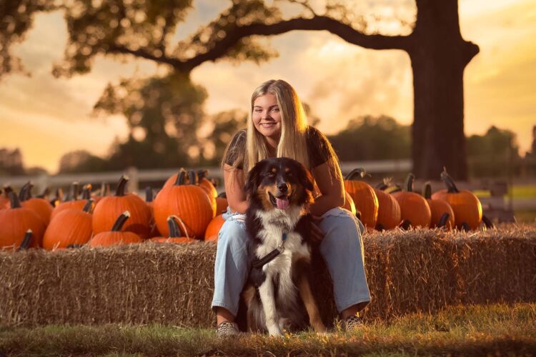 A girl and her dog sitting on hay bales in a pumpkin patch