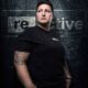 famale sports massage therapist with short hair and strong build stands in front of dark logo wall looking off camera.
