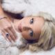 beautiful blonde model with blue eyes that match her sapphire jewelry, wearing white fur on a white bedspread.