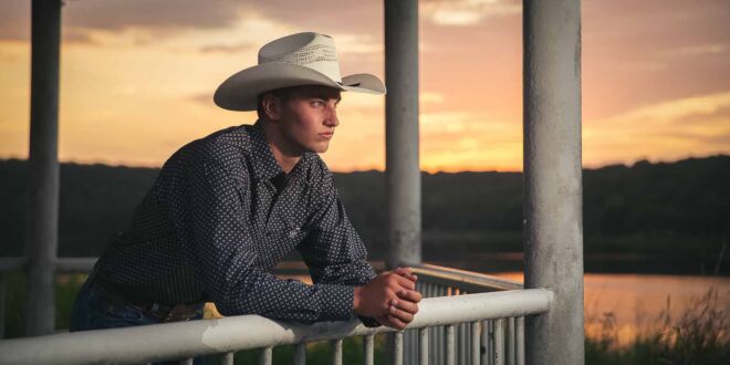 handsome young man in a cowboy hat looking out over a balcony on a lake at sunset