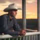 handsome young man in a cowboy hat looking out over a balcony on a lake at sunset