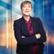 blonde short haired business woman laughs in front of a blue and orange urban window background, wearing a burgundy top and a blue blazer