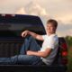 young man sitting in the back of his truck in a field at sunset